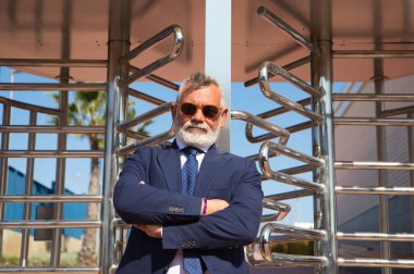 Handsome, mature, grey-haired man with beard, jacket and tie and sunglasses at the entrance to the office building where he works as chief accountant and executive. The man with his arms crossed.
