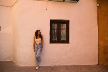 Gündelik giysiler içinde Seville 'deki eski bir evin beyaz duvarına yaslanmış genç ve güzel bir kadın. Kadın farklı ifadeler ve duruşlar sergiliyor ve tatilin tadını çıkarıyor..