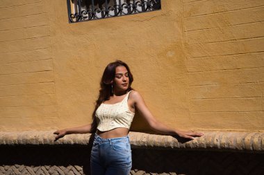 Seville 'de tatilde uzun kıvırcık saçlı güzel bir genç kadın. Model, kollarını açarak şehrin eski bir bölgesinde bir binaya yaslanarak poz veriyor. Tatil kavramı.
