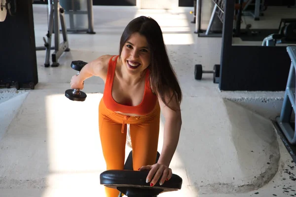 Beautiful young woman with a sculpted body exercising on a weight bench. She has one knee up and one foot on the floor and lifts a dumbbell. Concept Gymnastics, health and wellness.