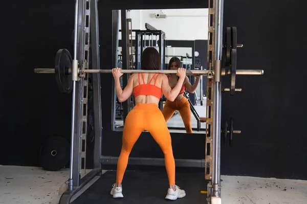 Young and beautiful woman with a sculpted body doing squat exercises with a dumbbell bar on her neck. The woman is wearing orange. Concept Gymnastics, health and wellness.