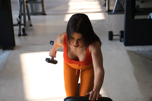 Beautiful young woman with a sculpted body exercising on a weight bench. She has one knee up and one foot on the floor and lifts a dumbbell. Concept Gymnastics, health and wellness.