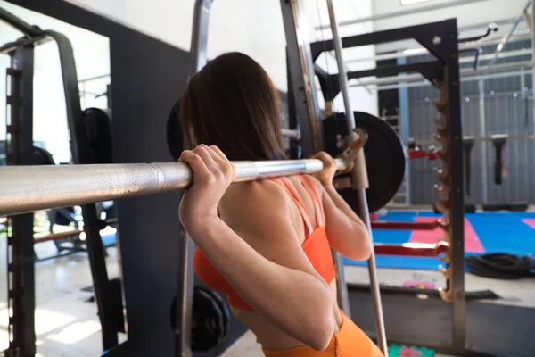 Young and beautiful woman with a sculpted body doing squat exercises with a dumbbell bar on her neck. The woman is wearing orange. Concept Gymnastics, health and wellness.