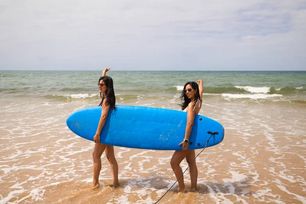 Two young and beautiful women with a surfboard on the shore of the beach. The women are enjoying their trip to the beach paradise. Holidays and travels.