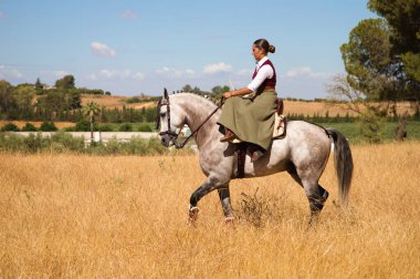 Young and beautiful Spanish woman on a Thoroughbred horse riding in the countryside in Spain. The woman is wearing a horse riding uniform. Thoroughbred and equine concept.