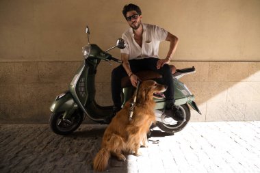 A handsome young man with his brown golden retriever dog. The man is sitting on his retro style motorbike and the dog is sitting on the ground. Concept pets, animals, dogs, pet love, golden retriever.