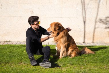 Sakallı ve gözlüklü yakışıklı genç adam çimenlerde oturmuş köpeğiyle güneşleniyor. Evcil hayvanlar, hayvanlar, köpekler, evcil hayvanlar, seyahat.
