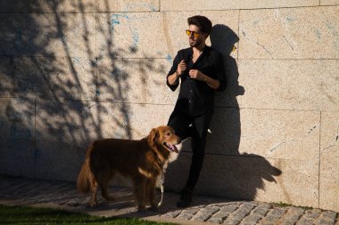 Sakallı ve gözlüklü yakışıklı genç adam kahverengi golden retriever köpeğiyle duvara yaslanıyor. Evcil hayvanlar, hayvanlar, köpekler, evcil hayvanlar, seyahat.