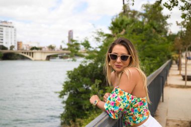Young, pretty blonde woman with sunglasses leaning on the railing of the river. The woman is happy and smiling. In the background you can see the river in seville.