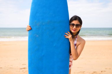 Mavi sörf tahtası olan genç ve güzel bir Latin kadın. Kadın kumsalda. Tatil ve yaz konsepti.