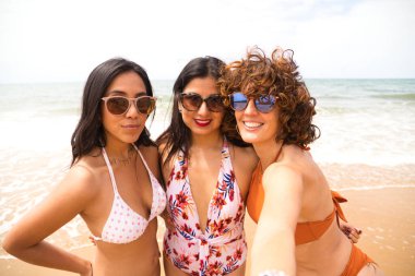 Sahilde arka planda deniz olan üç güzel kadın selfie çekiyor. Kadınlar tatildeler ve fotoğraf için gülümsüyorlar, güneş gözlüğü takıyorlar. Tatil ve seyahat kavramı.