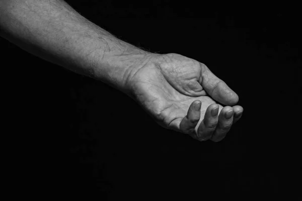 The outstretched wrinkled left hand of an elderly man asks for help on a black background. Black and white.