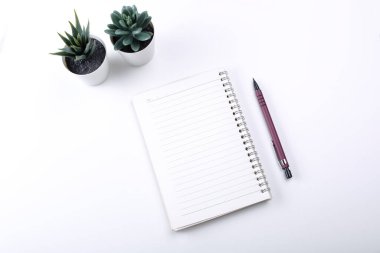 Boş defterli beyaz ofis masası. Not defteri, kalem, beyaz masadaki yeşil yapraklı çalışma alanı.
