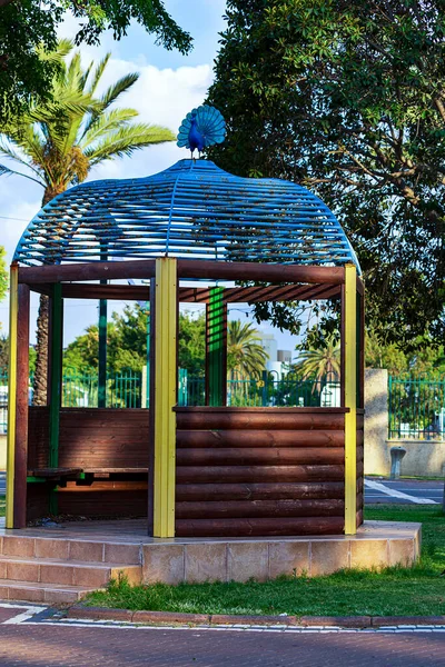 Bahçe çardağının tepesinde metal tavus kuşu. Rahat bahçe mobilyaları olan bir yazlık ev resmi. Mavi gökyüzü arka planında ahşap bir çardağı ve yeşil ağaçları olan güzel bir bahçe.. 