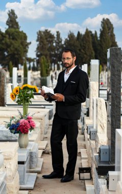 Siyah kippah içinde sakallı Yahudi İbrani İncili okuyup dua ediyor. İsrail 'de bir Yahudi mezarlığında dua etmek. Sığ bir alan derinliğine sahip Yahudi mezar taşları. Seçici odaklanma. Ormandaki eski Yahudi mezarlığı.