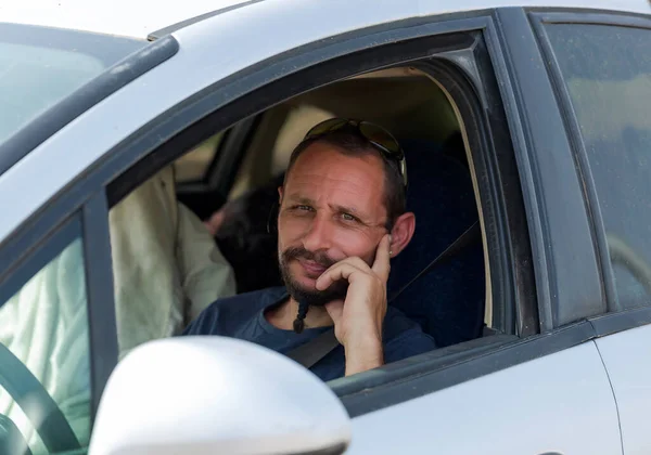 The beard caucasian man is alone driving a white car. Seen from the window of a male driver while behind the wheel. Young handsome man sitting in his automobile behind the wheel. Guy in car, close up