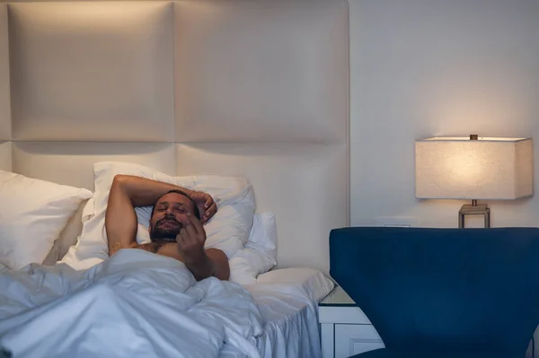 Naked bearded man beckoning someone to go to bed. Nude muscular handsome caucasian guy lying in bed on back under a white blanket with hand behind head, beckons with his finger, doing coming gesture