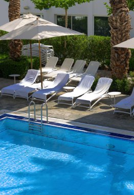 Açık havuzun etrafında şemsiyeler ve plaj sandalyeleri (güneş yatakları) ve gevşemek için hurma ağaçları vardır. Tatil ve yaz. Açık hava, mavi su ve havuza çıkan merdivenlerle muhteşem bir yüzme havuzu.