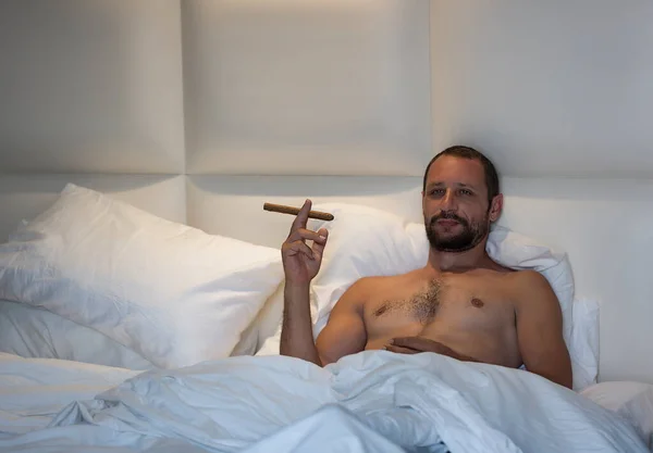 Handsome muscular man wants to smoke cigarette on his bed. Naked bearded caucasian guy lying in bed under a white blanket with wants to lighting cuban cigar. Good looking smoking man in bedroom. Rests