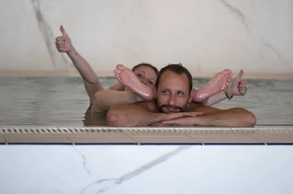 Laughing and leisure, couple enjoying relaxing in the swimming pool. Focus is on handsome muscular man. Female feet above water. Bearded man with has happy face. Her sexy legs on the man\'s head