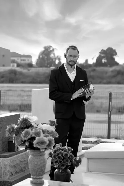 Black and white photo of  Bearded Jew in kippah reading a Hebrew Bible, praying. Prayer in jewish cemetery. Old jewish cemetery. Hasidic jew reading Torah. Vertical orientation. Selective focus