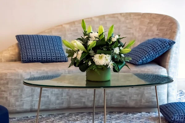 Living room decor: glass coffee table with white chrysanthemums, lilies bouquet and sofa on background. Home decoration. Beautiful luxury bouquet of mixed flowers in vase. Ikebana, flower arrangement