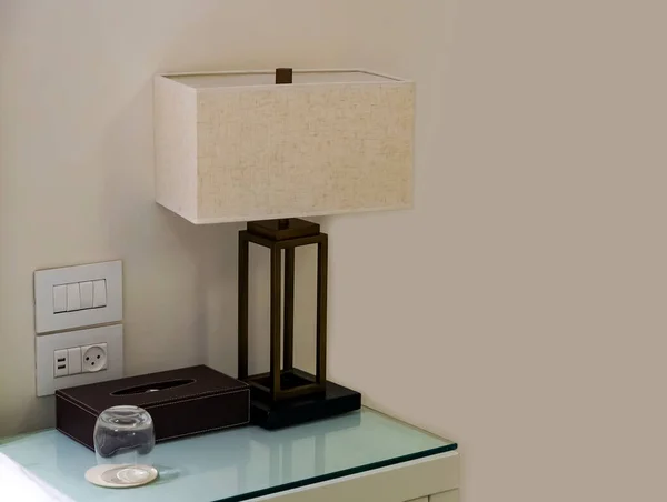 Modern bedroom lamp near bed with night table. Stylish lamp, paper napkins and glass of water on bedside table. Cozy atmosphere in the home interior, sleeping room or luxury hotel. Morning in bedroom