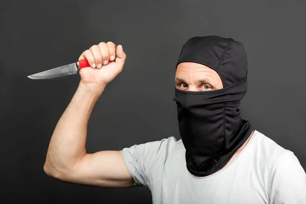 Man hidden in wearing black mask holding a pocket-knife and looking at the camera isolated on dark background. Robbery, violence and knife attack concept. Killer man attacks with a kitchen knife.