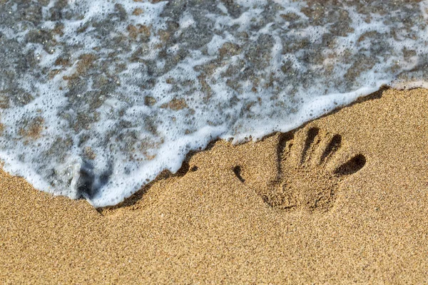 Human handprint at the sandy shore, transparent wave approaching. Palm print washing the waves of the sea. Closeup shot of handprints, nice nature summer background. Hand print on sea sand texture.