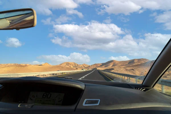Selective focus and front view through car\'s window on bendy road and mountain fantasy landscape on blue sky background with clouds. Sunny sky over cliffs, large salt mountains Judean desert, Israel