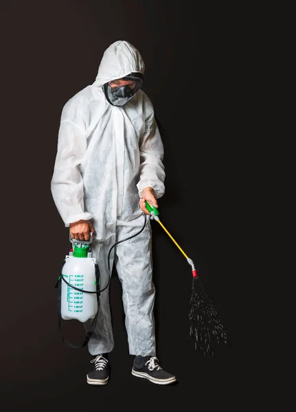 Male wearing protective clothing: white sterile uniform with facial mask, hand pressure pump disinfecting. Worker wearing protective suit using disinfectant manual sprayer, isolated on dark background