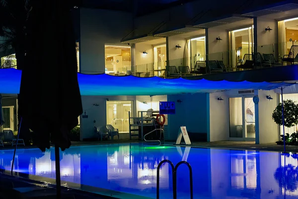 Blue luxurious swimming pool at night with modern house. Water of swimming pool illuminated in blue by twilight. Modern villa with colored led lights at night, lounge chairs, palm tree. Trendy design