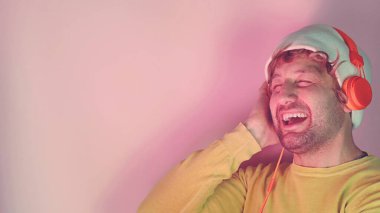 Neşeli, kaygısız sakallı adamın yatay çekimleri beyaz şapka takar, sarı gömlek giyer, yüksek sesle müzik dinlerken pembe arka planda izole edilmiş turuncu kulaklık takar..