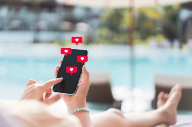 Akıllı telefon kullanan ve kalp sohbet ikonuyla yatakta oturan bir kadın. Yüzme havuzu tatil köyünde soyut bir arka plan. Teknoloji işi ve seyahat tatili konsepti. Klasik ton filtre efekti renk biçimi.