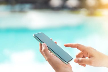 Kadın eli, tropik plaj arka planında akıllı telefon kullanıyor. Teknoloji işi ve seyahat tatili konsepti. Klasik ton filtre efekti renk biçimi.