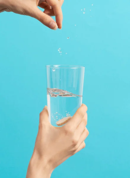 a female hand drops an effervescent tablet into the glass of water on the blue background