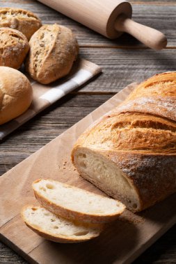 Doğrama tahtasına taze pişmiş ekmek, üzerinde merdane ve tam tahıl ruloları var. Ekmek ve ekipmanlar eski ahşap masada açıkta..