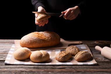 Youn Lady baker cuts her freshly beked bread on the chopping board. Bread and whole grains buns exposed onthe kitchen cloth placed on the wooden surface.