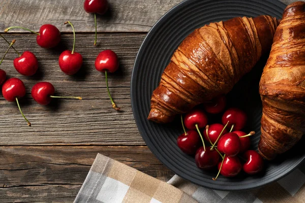 Freshly baked croissant exposed on the decorative platter with juicy cherries scattered on the side. Everytking placed on the vintage wooden table.