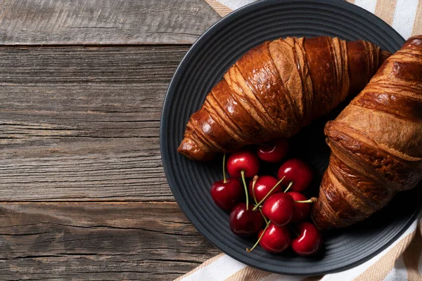 Freshly baked croissant exposed on the decorative platter with juicy cherries scattered on the side. Everytking placed on the vintage wooden table.