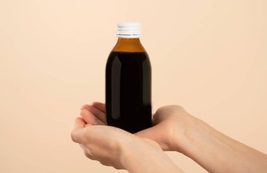 Kahverengi bir şişe soğuk algınlığı ilacı tutan eller. Ürün yerleştirme veya ürün sunumu için etiketsiz şişe. Bej arkaplan.