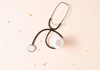 Top view of stethoscope, medication bottle and pills scattered all over the beige background. Medical equipment for measuring the heart rate, lungs, and abdomen sounds.