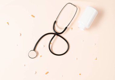 Top view of stethoscope, medication bottle and pills scattered all over the beige background. Medical equipment for measuring the heart rate, lungs, and abdomen sounds.