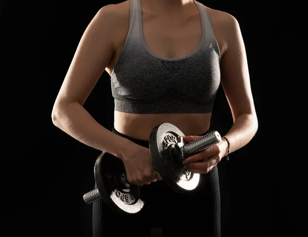 Close up image of slim and sporty woman with steel dumbbell in hand on the black background
