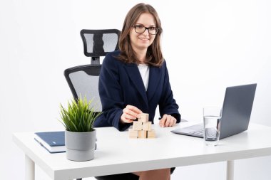 Young caucasian bussines woman stacking wooden blocks in her work office. Office interior, laptop, notebook and flower on white desk.