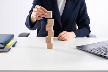 A close up image of bussines woman woman stacking wooden blocks in her work enviroment. Office interior laptop, glass of water and notebook on white desk.