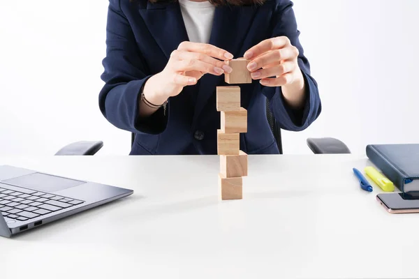 A close up image of bussines woman woman stacking wooden blocks in her work enviroment. Office interior laptop, glass of water and notebook on white desk.