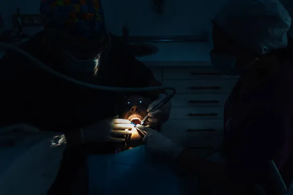 Dentistas que trabajan en un procedimiento dental en un paciente con la boca abierta bajo luz brillante en una habitación oscura. — Foto de Stock