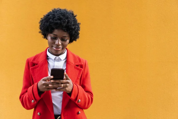 Mulher afro elegante digitando no telefone celular enquanto está ao ar livre contra uma parede amarela. — Fotografia de Stock