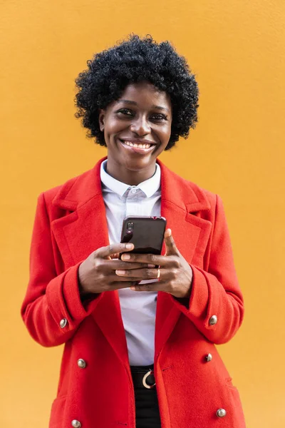 Mulher afro-americana olhando para a câmera e sorrindo enquanto segurava um telefone celular. — Fotografia de Stock